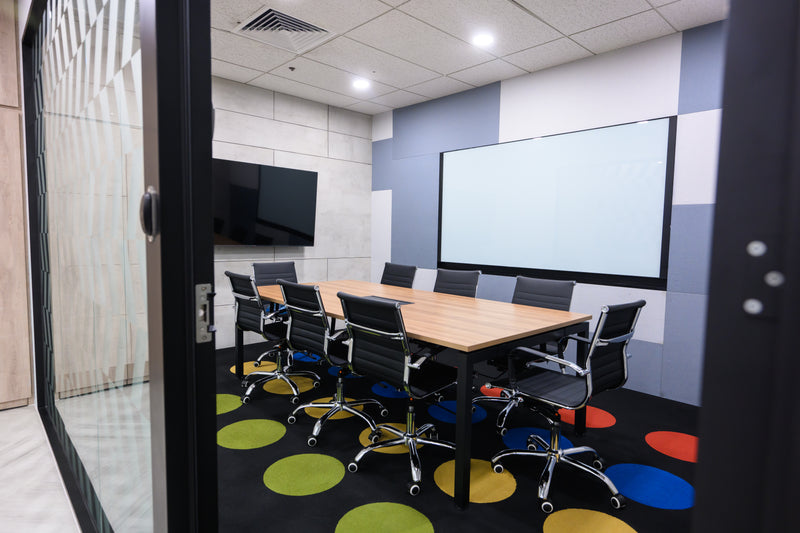 This is a photo of our sizable meeting room featuring an LG TV with wireless casting capabilities. The meeting room table is encircled by eight computer chairs. The floor is polka dotted. A glass whiteboard is also embedded on the wall.